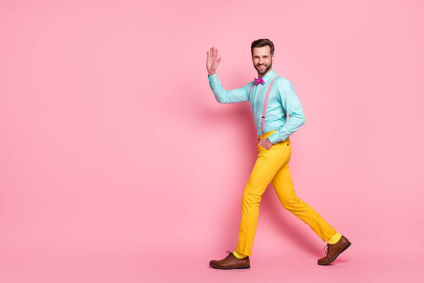 Full size profile photo of handsome guy trend stylish look walk red carpet celebrity waving hand wear shirt suspenders bow tie yellow pants footwear isolated pastel pink color background - 写真・画像