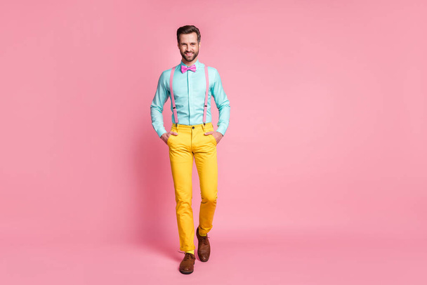 Full size photo of handsome guy trend clothes beaming smile walking office meeting wear teal shirt suspenders bow tie yellow trousers boots socks isolated pastel pink color background - 写真・画像
