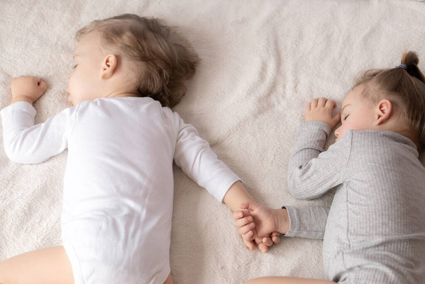 Gyerekkor, alvás, pihenés, család, életmód koncepció. két 2 és 3 éves, fehér és bézs testruhába öltözött kisgyermek bézs és fehér ágyon alszik ebédnél, kéz a kézben, felülnézetben. - Fotó, kép