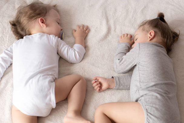 Childhood, sleep, relaxation, family, lifestyle concept. two young children 2 and 3 years old dressed in white and beige bodysuit sleep on a beige and white bed at lunch holding hands top view - Fotoğraf, Görsel
