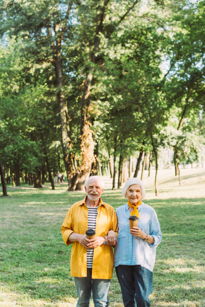 Весела літня пара з паперовими чашками дивиться на камеру в парку
  - Фото, зображення