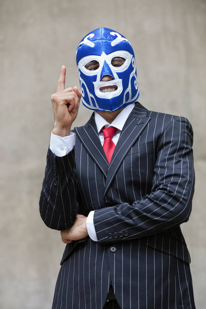 Young businessman in pinstripes suit and wrestling mask pointing up over gray background - Fotografie, Obrázek