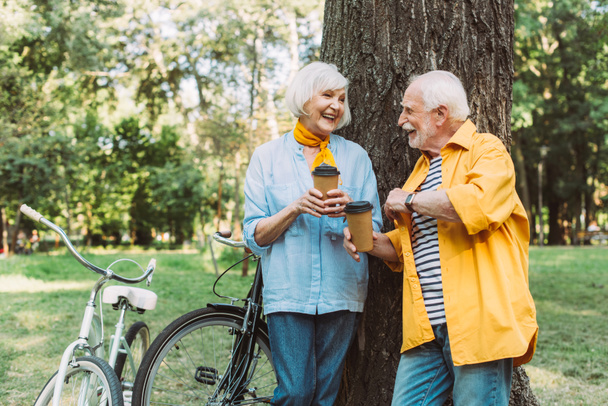 Selective focus of elderly couple with paper cups smiling at each other near bikes in park   - Photo, image