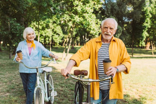 Selective focus of smiling senior man holding paper cup near wife with bike in park  - Фото, зображення
