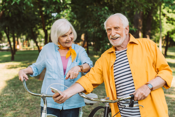 Mosolygó idős nő ujjal mutogatott férj közelében okostelefon és kerékpárok a parkban  - Fotó, kép