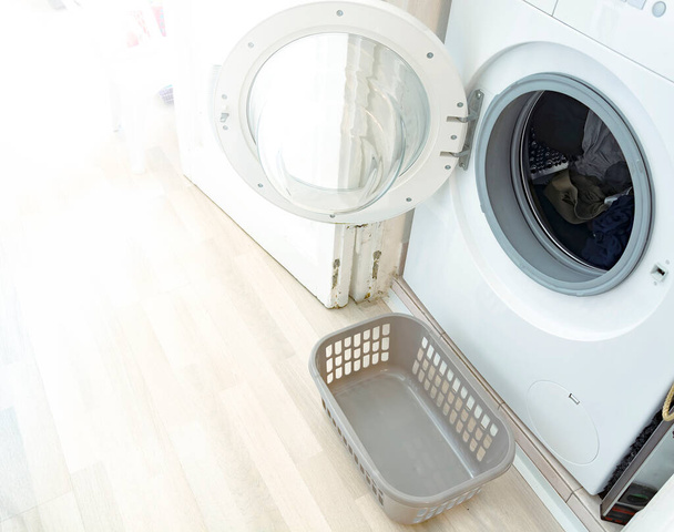 egy nyitott ajtós mosógép és egy kis műanyag kosár a frissen mosott szennyes tárolására. Egy ház belseje. Házimunka és ruhatisztítás. - Fotó, kép