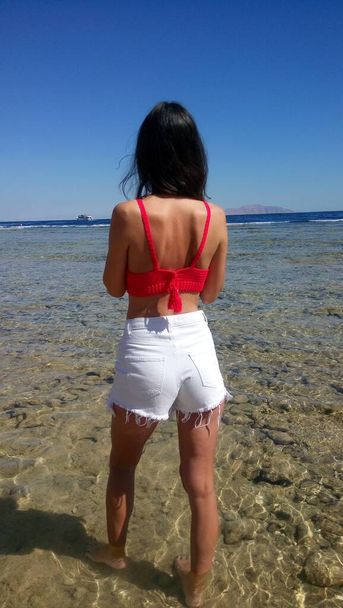 白いショートパンツの女の子と海の背景に赤いトップ - 写真・画像