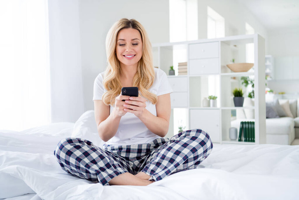 Photo of pretty house wife lady hold telephone writing blog social network post sitting cosy bed freelancer quarantine white linen legs crossed perfect sunny morning wear pajama room indoors - Photo, image