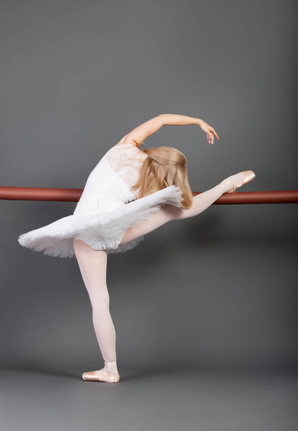 Young female ballet dancer stretching at ballet bar over grey background - Фото, изображение