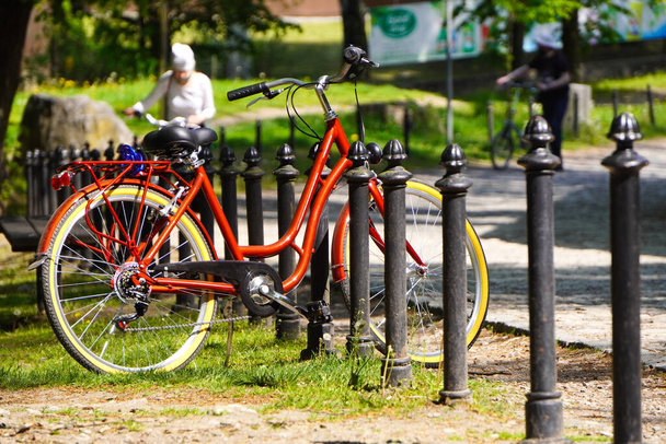 Parlak kırmızı bisiklet, güneşli bir günde yeşil bir parkın çitlerinde duruyor. Her gün bisikletle taşınıyorum. Bisiklet dışarıdaki park yerinde. Şehirdeki çevre dostu ve spor ulaşım araçlarının taşınması - Fotoğraf, Görsel