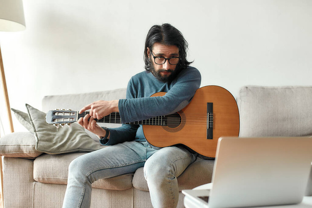 Young caucasian man sitting on sofa at home and adjusting acoustic guitar, watching guitar lesson online on laptop - Фото, зображення