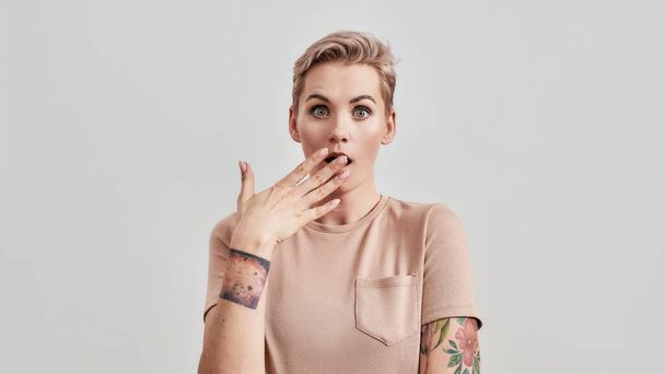 Portrait of a young attractive tattooed woman with pierced nose and short hair in beige t shirt looking surprised at camera with hand over her mouth isolated over light background - Photo, Image
