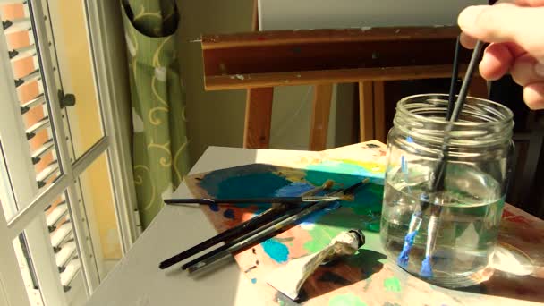 Hombre pintando con pintura al óleo y limpiando sus pinceles en un frasco con agua junto a una ventana brillante. Materiales para pintar en casa: lienzo, paleta, caballete
.. - Metraje, vídeo