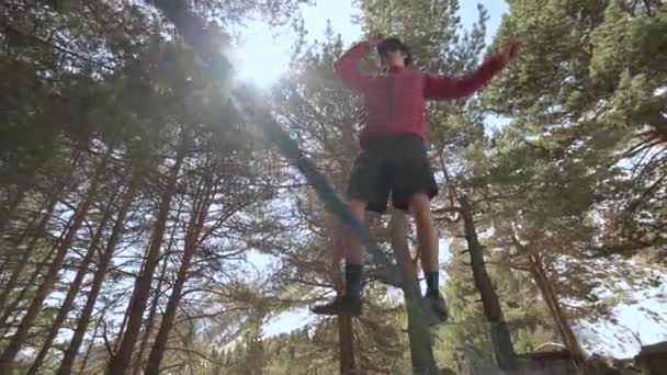 A young guy in a shorts jacket and a cap with sunglasses goes balancing on a stretched slackline in the coniferous forest in the afternoon. Outdoor sports and leisure concept - Materiał filmowy, wideo