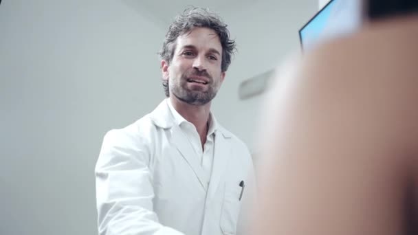 Professional medical appointment with male doctor in medical centre, young physician in white lab uniform greeting female patient and inviting to the office for medical consultation. Health care and - Πλάνα, βίντεο