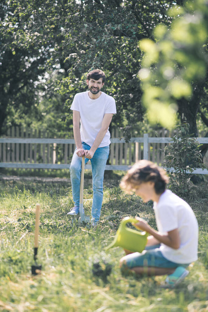 Dark-haired boy watering the plant, his father watching him and smiling - Fotoğraf, Görsel