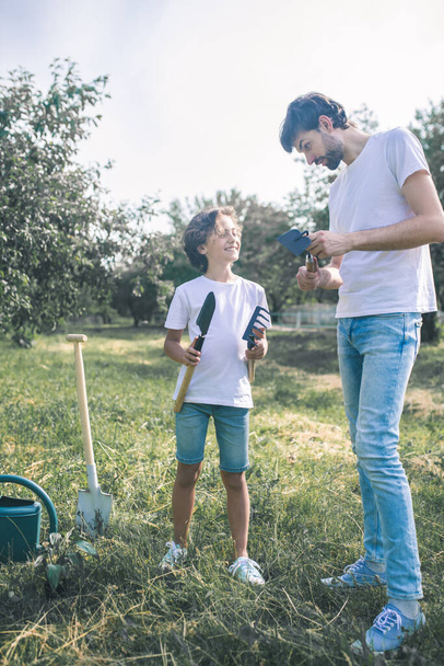 Dark-haired boy and his dad holding gardening equipment and looking ready to work - Photo, image