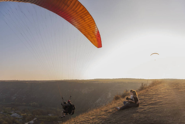 A happy family with a dog are sitting on the top of the hill, watching the paragliders fly in the setting sun. A young man embraces a beautiful girl who holds a Schnauzer in her arms. Traveling with pets on the weekend - Foto, imagen