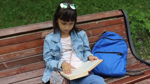 meisje schoolmeisje met een rugzak zit op een park bank en leest een boek - Video