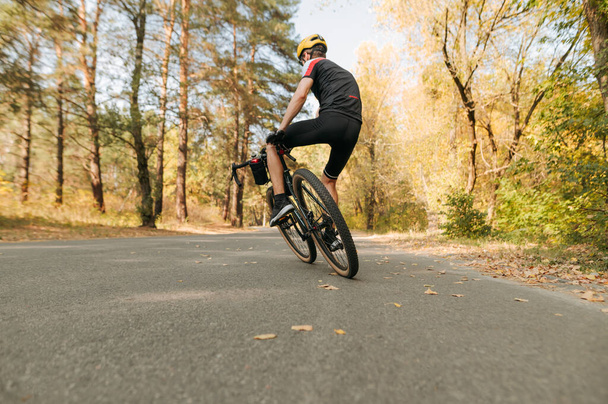 自転車競技者は道路上の秋の公園に乗り、自転車でトリックを行い、自転車を側面に置きます。街の外の秋の道を自転車で歩く男、バックビュー. - 写真・画像