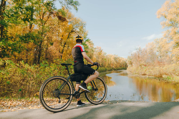 Spor kıyafetli profesyonel bisikletçi güzel bir sonbahar manzarasında parktaki bir gölün kıyısında duruyor. Bisikletçi bisiklete biner ve güzel sonbahar manzarasına bakarak rahatlar.. - Fotoğraf, Görsel