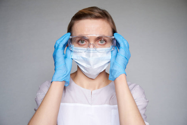 Close up of female doctor or scientist with a medical mask and hands in latex gloves over grey background. She is putting on glasses - Photo, Image
