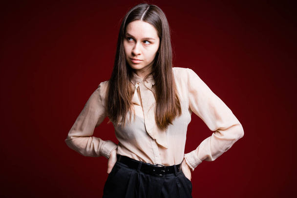 ライトシャツを着たブルネットは彼女のベルトを手に取り、側面に目を向ける。若いです女性でオフィスの服で赤の背景. - 写真・画像