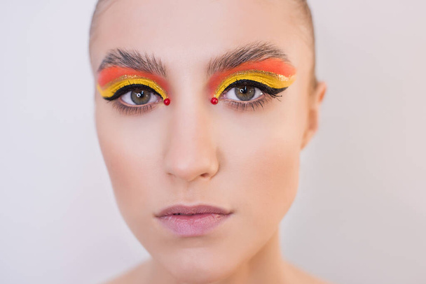 Close-up portrait of a fashion girl with bright makeup. Orange-yellow shadows on the eyes, black eyeliner, red beads near the eyes, pink lips, laminated eyebrows, clean face, hair gathered in a bun on a gray background. - 写真・画像