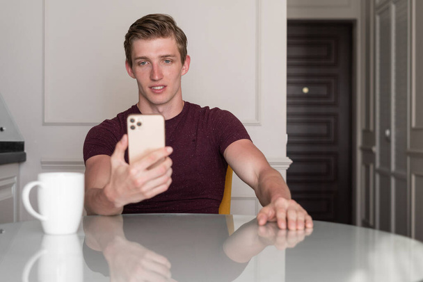 Attractive caucasian young man in trendy shirt texting messages via social networks and browsing internet, using wifi on his electronic device during coffee break at home - Valokuva, kuva