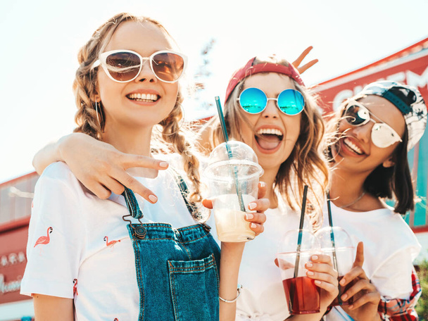 Three young beautiful smiling hipster girls in trendy summer clothes.Sexy carefree women posing outdoors.Positive models holding and drinking fresh cocktail smoothie drink in plastic cup with straw - Foto, afbeelding