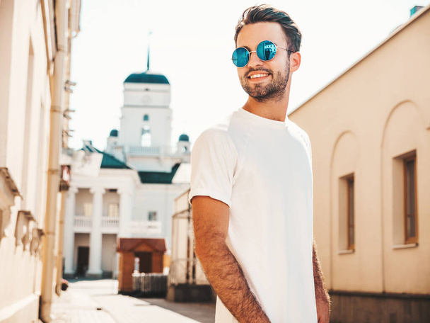 Portrait of handsome confident stylish hipster lambersexual model.Man dressed in white T-shirt. Fashion male posing on the street background in round sunglasses - Zdjęcie, obraz