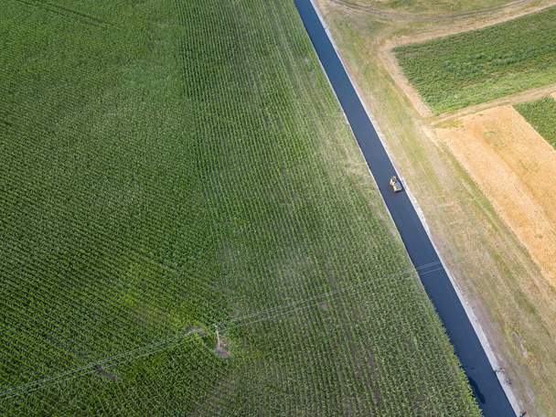 空中ドローンビュー。ウクライナの田舎の緑のトウモロコシ畑の隣にアスファルト道路の建設. - 写真・画像