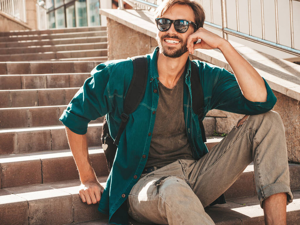 Portrait of handsome smiling stylish hipster lambersexual model.Man dressed in green shirt. Fashion male sitting at the stairs on the street background in sunglasses - Fotó, kép