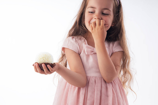 Красивая маленькая девочка удивлена вкусной шоколадной кокосовой конфеткой в виде мяча. Ребенок готов съесть торт.  - Фото, изображение