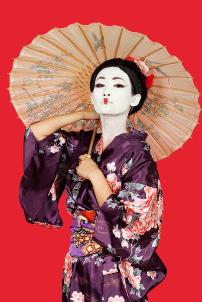 Portrait of Japanese woman in kimono holding parasol against red background - Foto, imagen