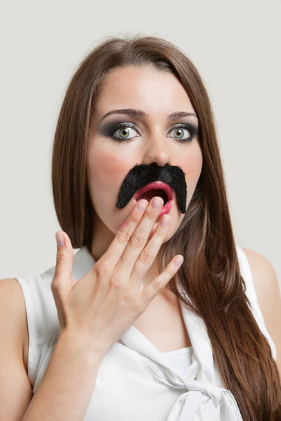 Portrait of shocked young woman with fake moustache over gray background - Fotografie, Obrázek