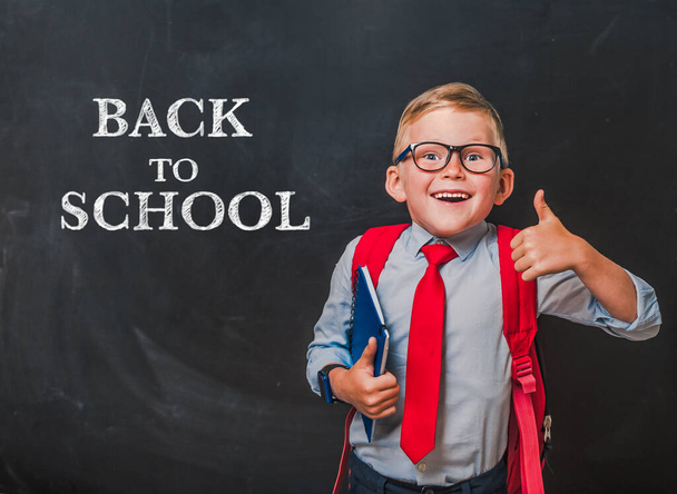Happy smiling boy in uniform with book and bag. Surprised kid in glasses go back to school. Success, motivation, winner, genius concept. Businessman against blackboard background. - Photo, Image