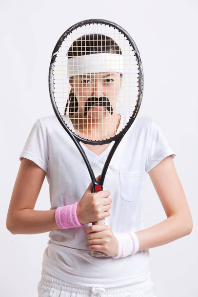 Portrait of young woman with a fake mustache holding tennis racket in front of her face against white background - Foto, afbeelding