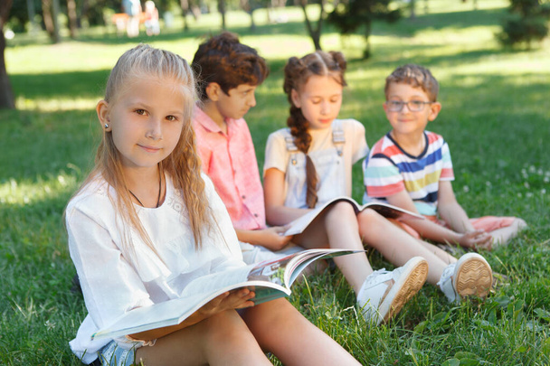Schönes kleines Mädchen liest im Park ein Buch, ihre Freunde ruhen sich im Gras aus - Foto, Bild