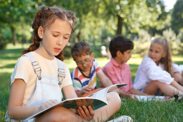 Young girl reading a book at the park, her friends relaxing on the grass on background - Photo, Image