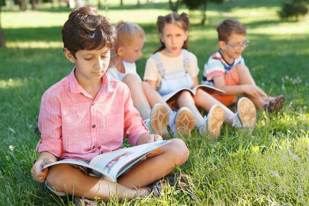 Kleiner Junge liest ein Buch, sitzt auf dem Gras im Park, seine Freunde reden im Hintergrund - Foto, Bild