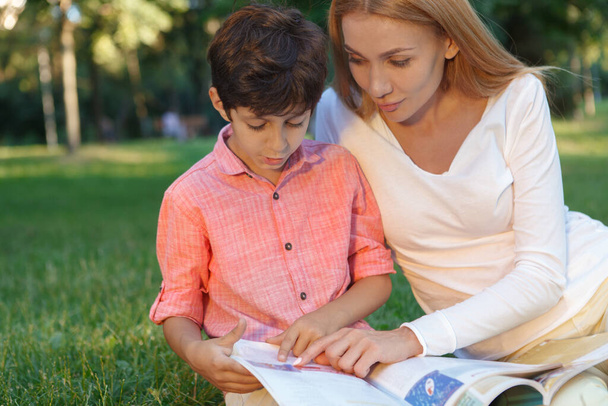 Close up of a cute little boy and his female teacher reading a book outdoors, copy space - Photo, Image