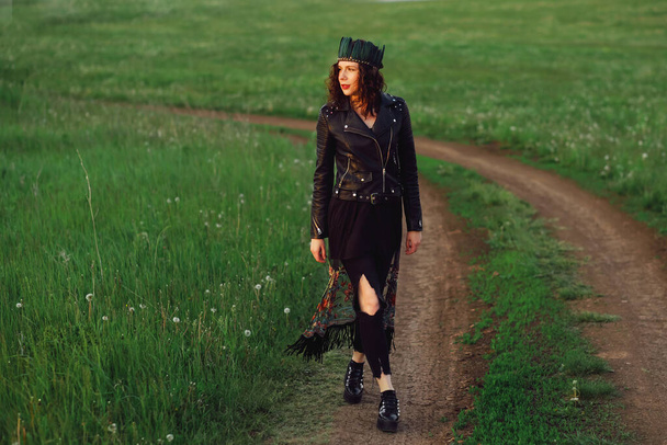 A young woman of 25-30 years old with curly red hair, in a black leather jacket and a tiara with feathers walks along a country road. - Фото, изображение