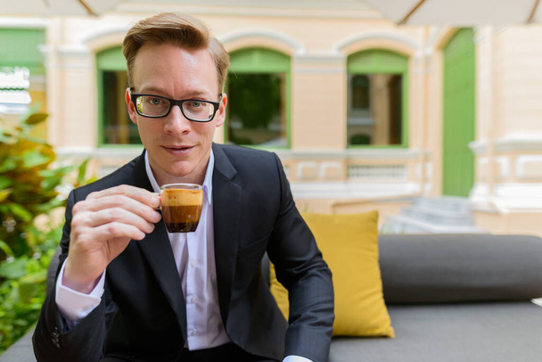 Portrait of young handsome blond businessman in suit relaxing at the coffee shop outdoors - Fotoğraf, Görsel