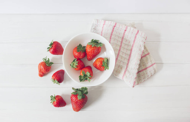 Top view of fresh strawberries on a white rustic wooden table, inside a bowl ready to eat as organic and healthy food, next to a cloth with red stripes  - Фото, зображення