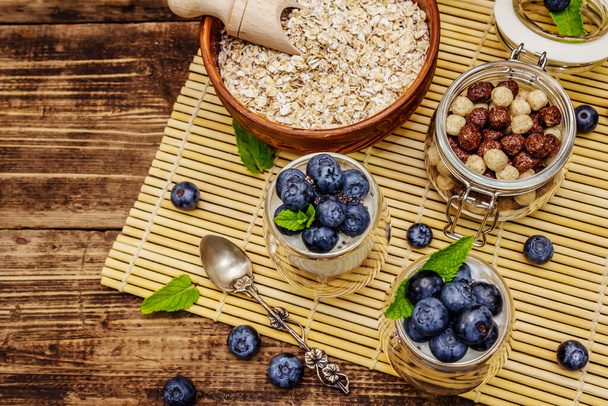 Healthy and useful breakfast concept. Yogurt with blueberries, oat-flakes and chia seeds. Ripe berries, mint leaves, silver spoon on wooden boards background - 写真・画像