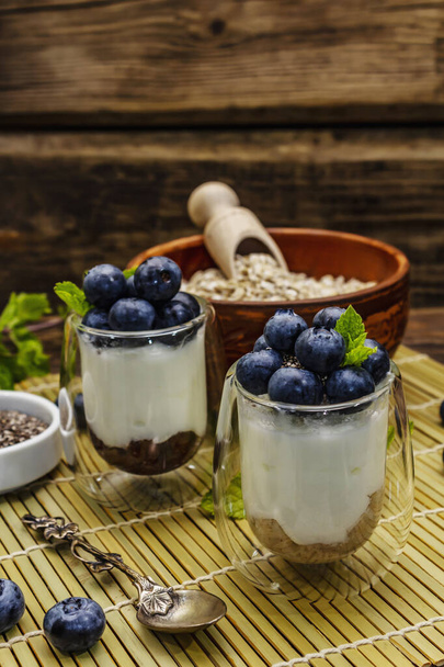 Gezond en nuttig ontbijt concept. Yoghurt met bosbessen, havervlokken en chia zaden. Rijpe bessen, muntblad, zilveren lepel op houten planken ondergrond, close up - Foto, afbeelding