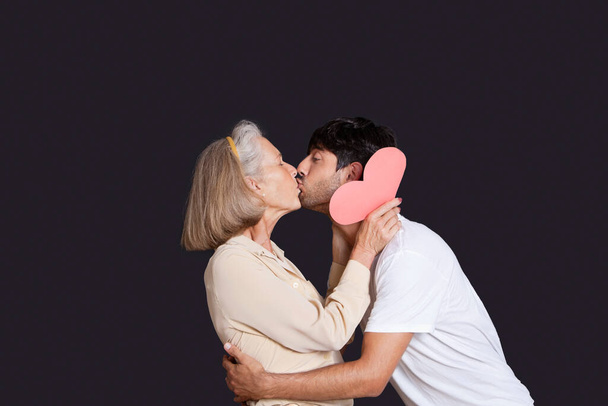 Young man kissing senior woman with red paper heart against black background - Fotó, kép