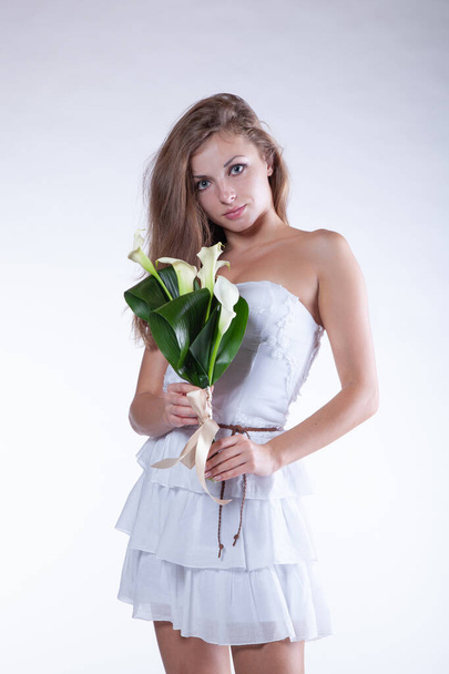 portrait d'une fille en vêtements blancs avec des fleurs dans le studio, se tient près d'un mur blanc - Photo, image