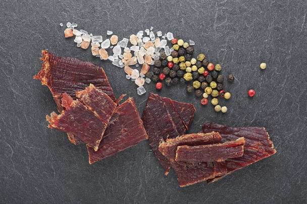 Beef jerky with peppercorns and salt on black stone table. Top view. - Фото, изображение